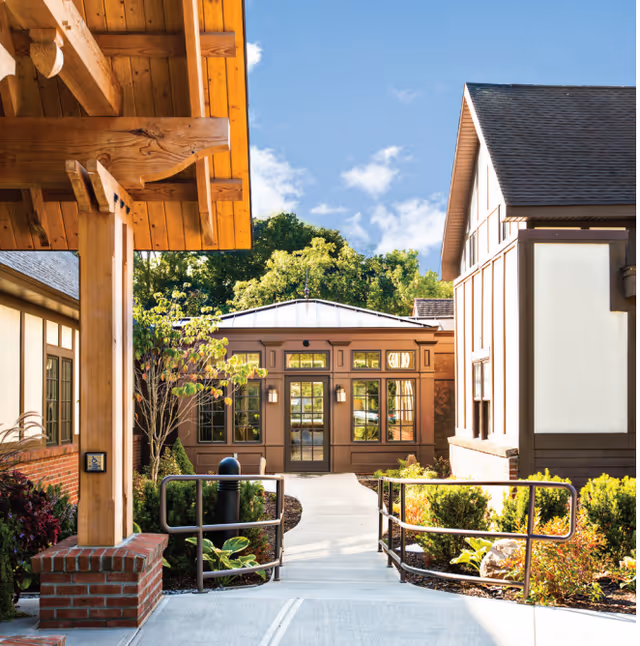 Sunlit exterior courtyard with a paved walkway leading to a building entrance flanked by landscaped beds and timber and brick architectural details.