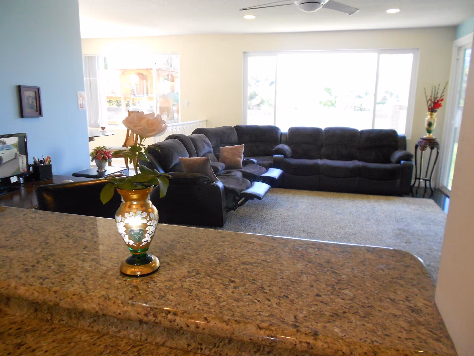 Interior view of a senior living facility's common area featuring a large dark sectional sofa with recliners, a carpeted floor, and large windows letting in natural light. In the foreground, there is a granite countertop with a decorative vase holding a single white rose. The room has light-colored walls and a ceiling fan.