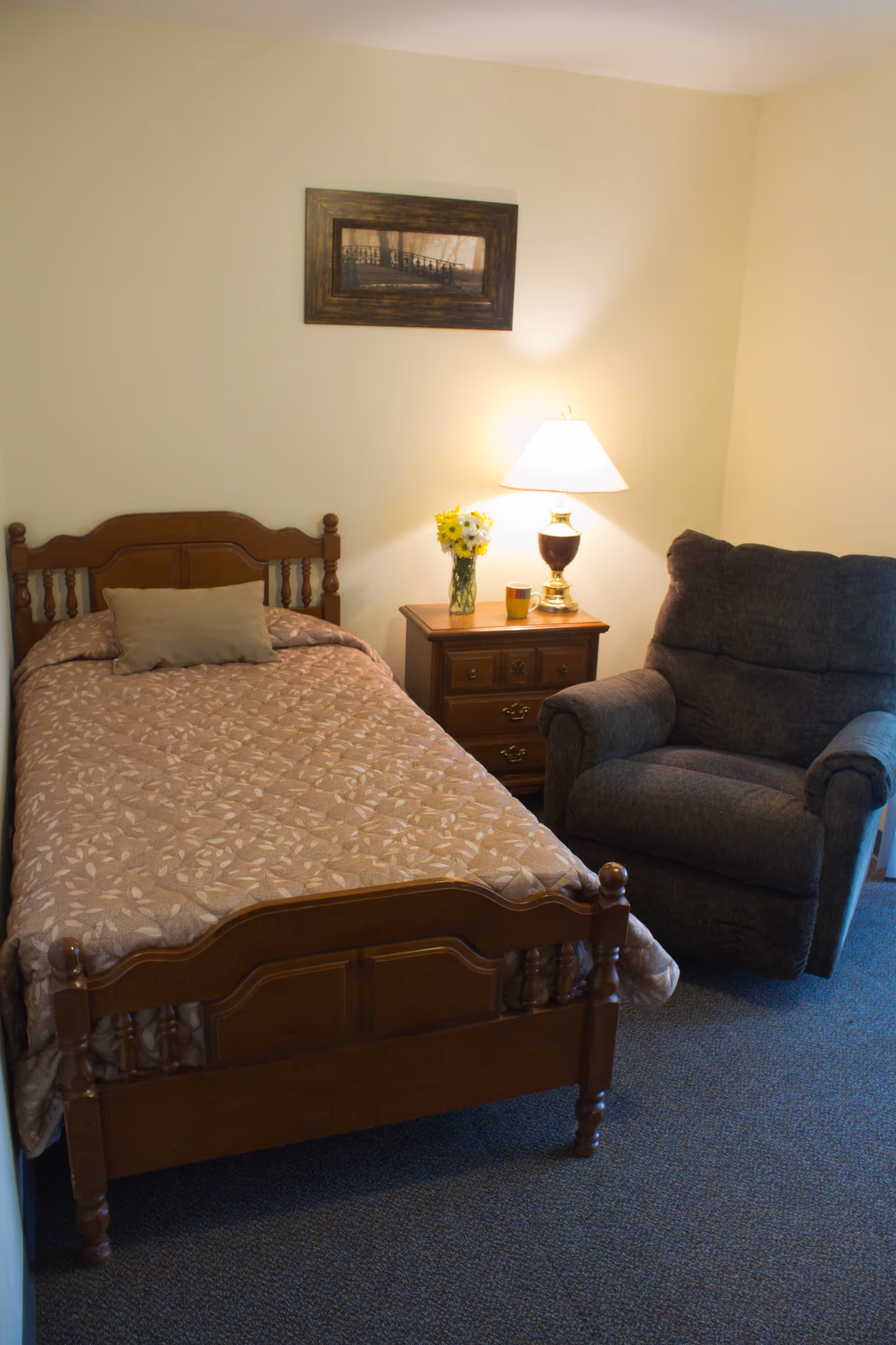 A small bedroom with a single wooden bed covered with a patterned beige bedspread and a beige pillow. Next to the bed is a wooden nightstand with a vase of yellow and white flowers, a coffee mug, and a lit table lamp. To the right of the nightstand is a dark gray upholstered armchair. A framed picture hangs on the wall above the bed.