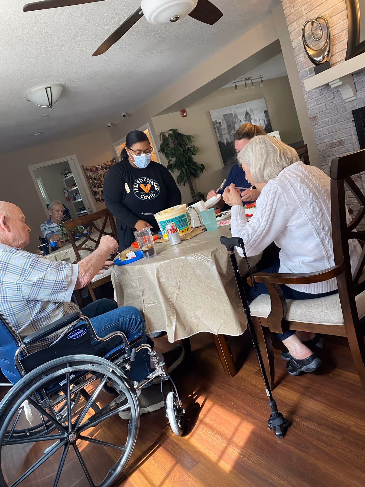 A group of elderly people and a caregiver wearing a mask are gathered around a dining table in a well-lit room. One elderly man in a wheelchair and an elderly woman with a cane are seated at the table, while the caregiver stands nearby serving or assisting. The room has wooden flooring, a ceiling fan, and a decorative fireplace with a modern sculpture on the mantel.