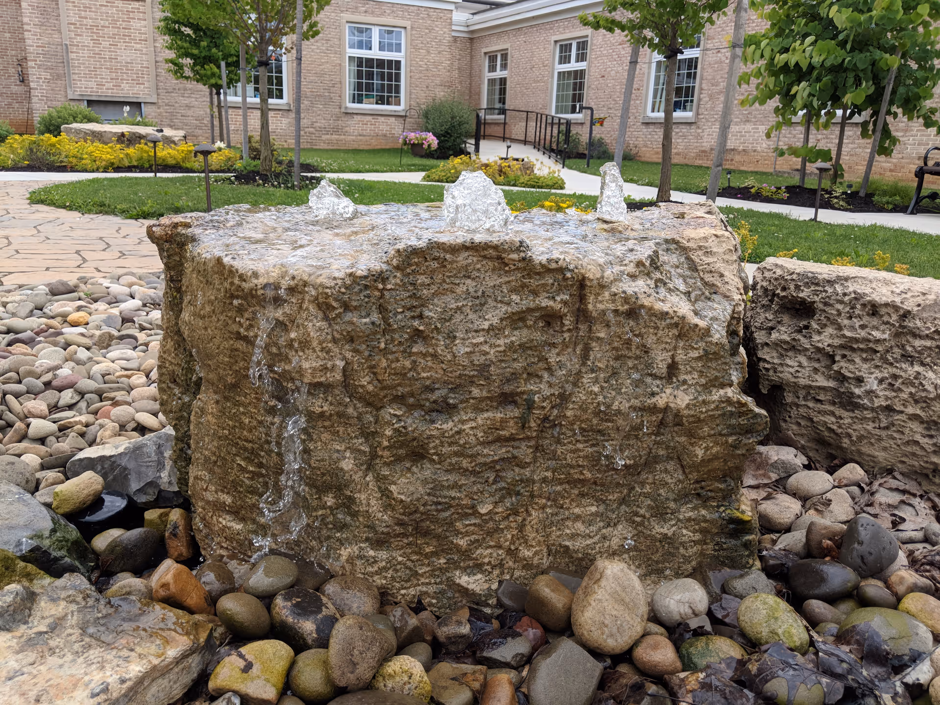 A stone water fountain with water bubbling up from three points on top, surrounded by smooth river rocks. In the background, there is a well-maintained garden with green grass, small trees, yellow flowers, and a brick building with windows and a ramp.