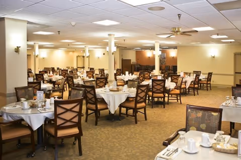 A spacious dining room with multiple round tables covered with white tablecloths, each set with plates, cups, and silverware. The room has beige walls, carpeted floors, ceiling lights, and ceiling fans. Chairs with cushioned seats and backs surround the tables.