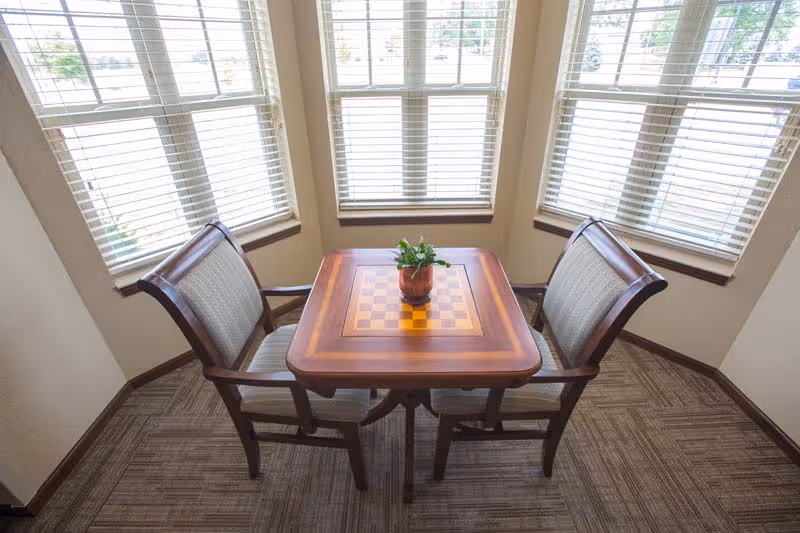 A small wooden table with a built-in chessboard pattern on the surface, placed in a corner surrounded by three large windows with white blinds. Two cushioned wooden chairs are positioned on opposite sides of the table, and a small potted plant is centered on the table. The floor is carpeted with a patterned design.