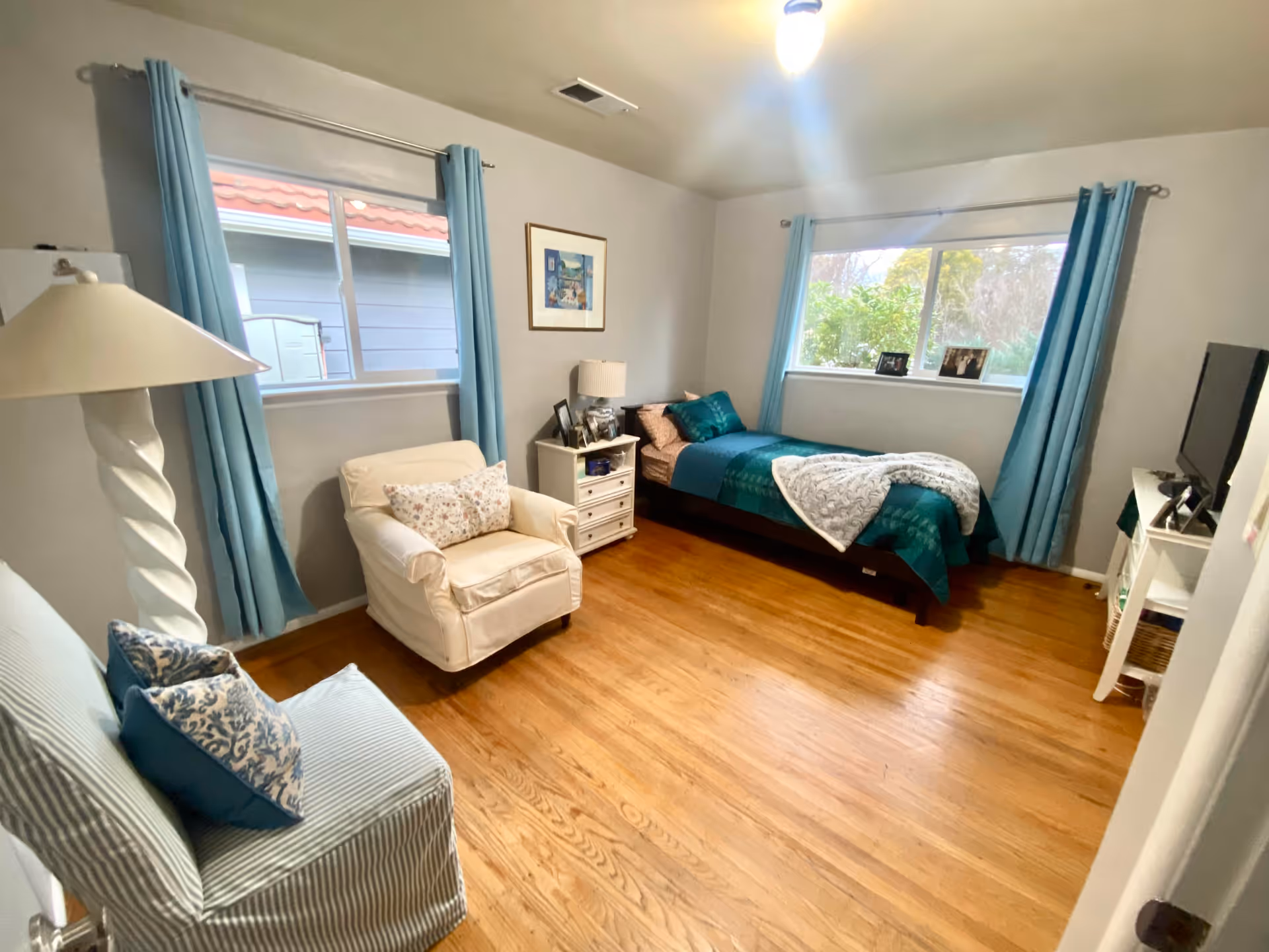 A cozy bedroom with wooden flooring, a single bed with teal and beige bedding, two windows with light blue curtains, a white armchair with a floral pillow, a striped chair with blue pillows, a white nightstand with a lamp and framed photos, and a white TV stand with a television.