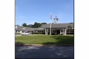 Single-story senior living building with a covered entrance, flagpole on a grassy lawn and a parked vehicle under a clear blue sky.