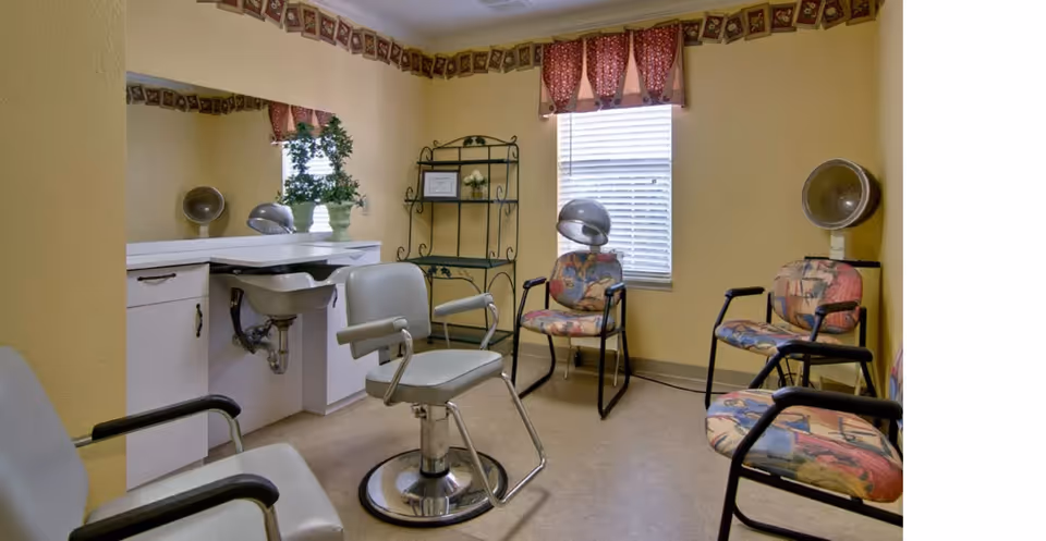 A small hair salon room with yellow walls, featuring a central salon chair in front of a counter with a sink and mirror. There are three additional chairs with colorful patterned cushions, two hair dryers mounted on stands, a window with blinds and a red valance, and a decorative metal shelf with plants and framed pictures.