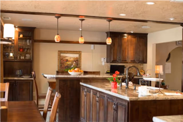A warm and inviting kitchen area with dark wooden cabinets and a large island countertop. The island has a sink and a small red potted plant on it. Three pendant lights hang from the ceiling above the island. In the background, there is a dining table with chairs and a framed painting on the wall.