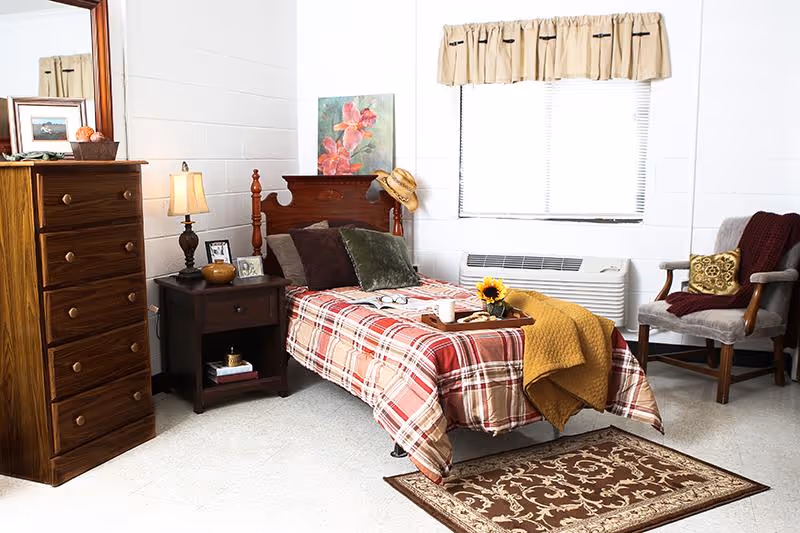 A cozy bedroom in an assisted living facility featuring a single bed with a red and beige plaid bedspread, a yellow throw blanket, and a tray with a sunflower and a cup. Next to the bed is a dark wooden nightstand with a lamp, framed photos, and a small bowl. A tall wooden dresser with a mirror stands to the left. There is a window with beige curtains above a heating/cooling unit, and a comfortable armchair with a burgundy throw and decorative pillow is placed near the window. A patterned rug lies on the floor in front of the bed.