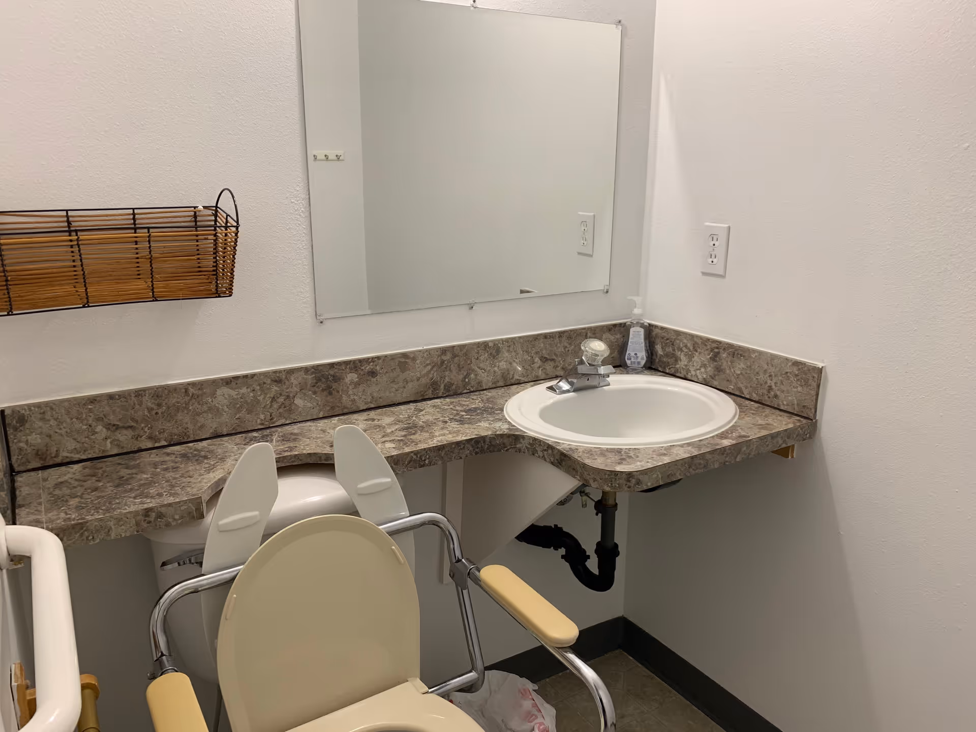 A small bathroom with a beige toilet equipped with armrests, a marble-patterned countertop with a white sink, a soap dispenser, a large rectangular mirror above the sink, a wall-mounted basket, and a white wall with an electrical outlet.