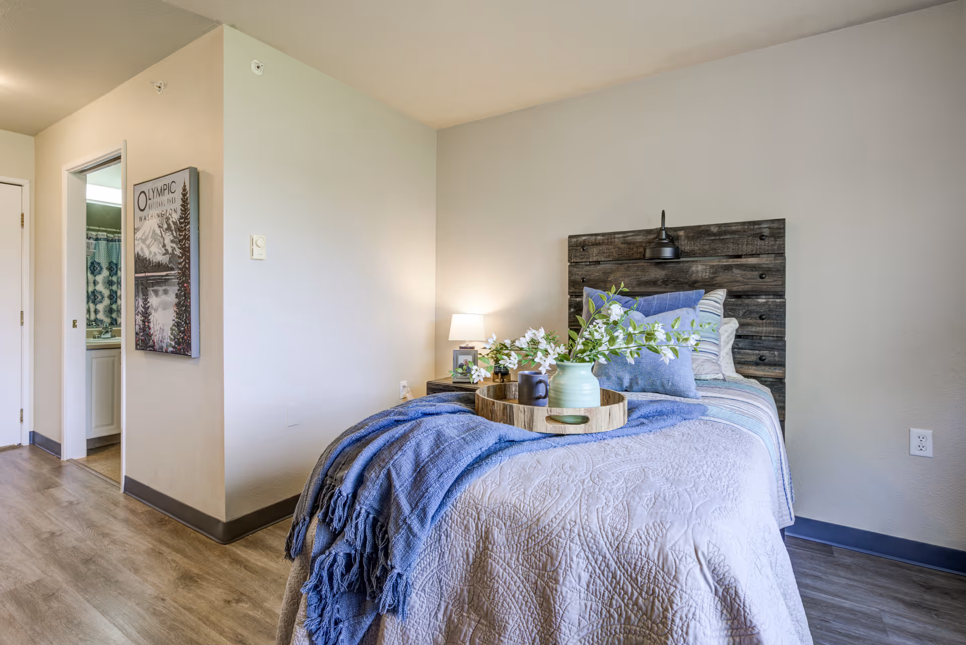 A cozy bedroom with a single bed featuring a rustic wooden headboard, decorated with multiple pillows and a blue throw blanket. A wooden tray with a vase of flowers and a cup is placed on the bed. To the left, there is a nightstand with a lamp and a framed photo. The room has light-colored walls and wood flooring. An open doorway reveals a bathroom with a shower curtain visible.