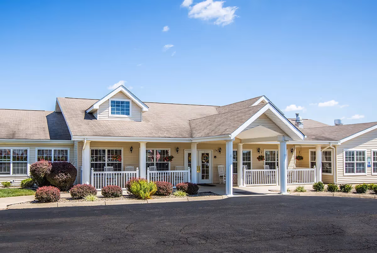 Exterior view of a single-story senior living facility building with beige siding, white trim, and a covered entrance supported by white columns. There are several windows, hanging flower baskets, and a paved driveway in front. The sky is clear and blue.