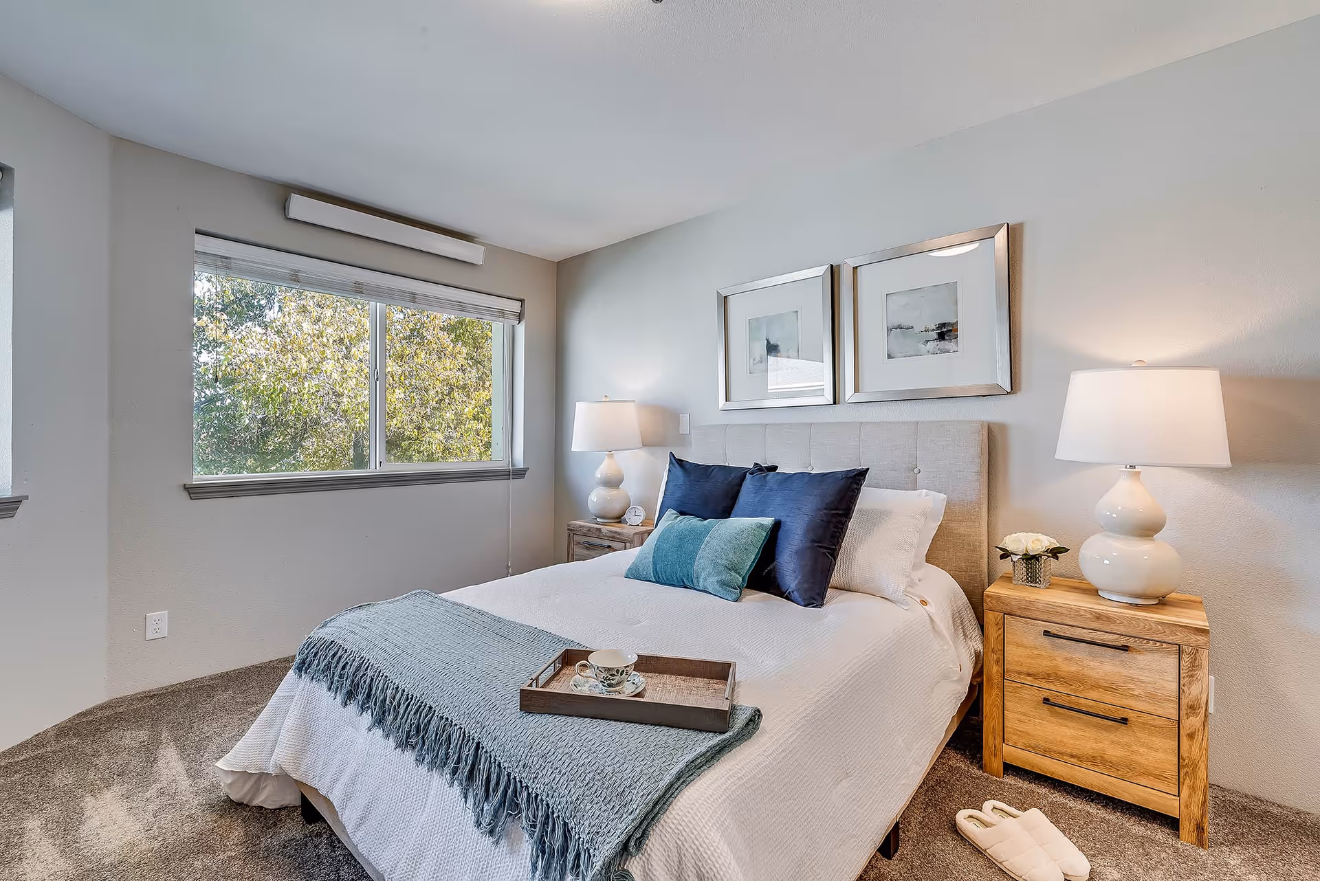 Bright furnished bedroom with a bed, pillows, bedside tables, lamps, and a window showing trees outside.