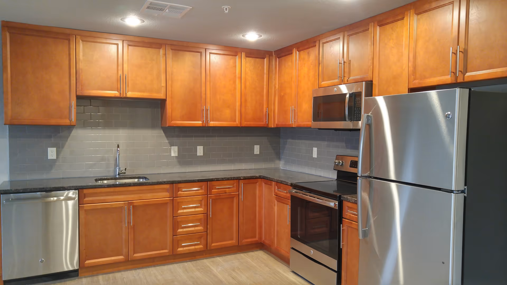Modern kitchen with wooden cabinets, gray subway tile backsplash, stainless steel appliances including a refrigerator, microwave, oven, and dishwasher, and a black countertop with a sink.