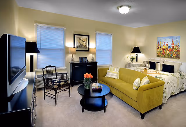A cozy senior living room and bedroom area with a yellow sofa, a round black coffee table with a vase of orange tulips, a wooden chair with floral cushion, a large bed with floral bedding and multiple pillows, two windows with blinds, a black cabinet with lamps and books, a flat-screen TV on a black dresser, and colorful floral artwork on the wall.