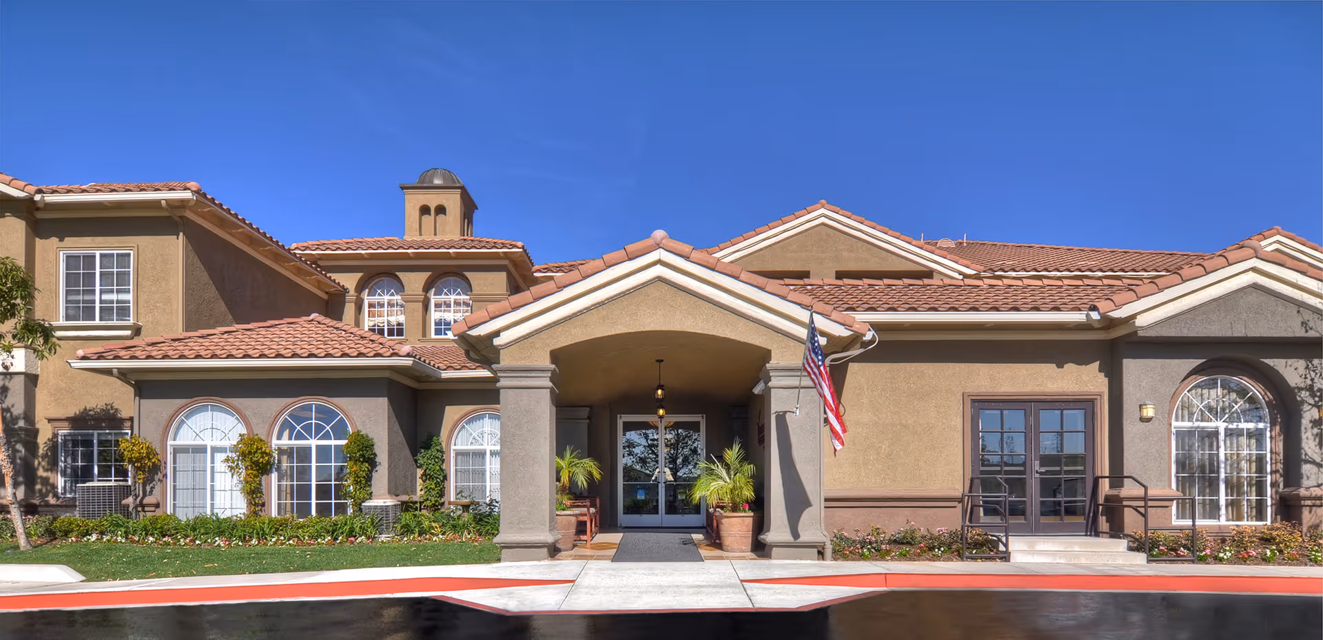 Front exterior view of a senior living facility building with a covered entrance, potted plants, an American flag, and multiple windows under a tiled roof on a sunny day.