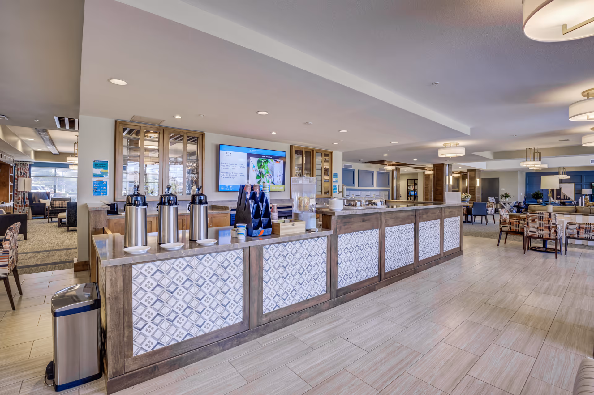 A spacious and well-lit common area in a senior living facility featuring a long counter with coffee dispensers, cups, and a water dispenser. Behind the counter are cabinets with glass doors and a large flat-screen TV mounted on the wall displaying information. The area includes comfortable seating with patterned chairs and tables, modern light fixtures on the ceiling, and a clean tiled floor.