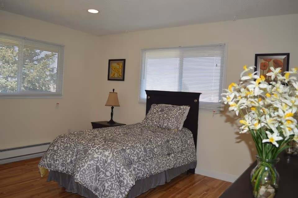 Bright bedroom with a single bed covered in gray patterned bedding, a nightstand and lamp, windows with blinds, and a vase of white and yellow flowers.