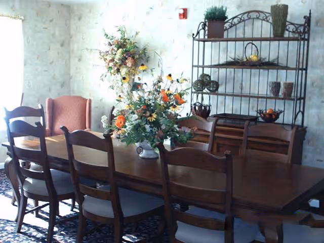 A dining room with a long wooden table surrounded by eight chairs. A large floral arrangement is placed in the center of the table. In the background, there is a metal shelving unit with decorative items and a red upholstered armchair near a window with sheer curtains.