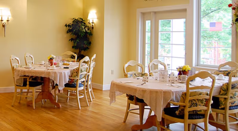 A bright dining room with two round tables covered with white tablecloths, each surrounded by six wooden chairs with cushions. The tables are set with cups, plates, and small flower arrangements. There is a large window letting in natural light and a potted plant in the corner. The walls are painted light yellow and there are wall-mounted lamps providing additional lighting.
