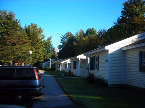 Row of single-story white apartment buildings along a sidewalk with a parked car and trees.