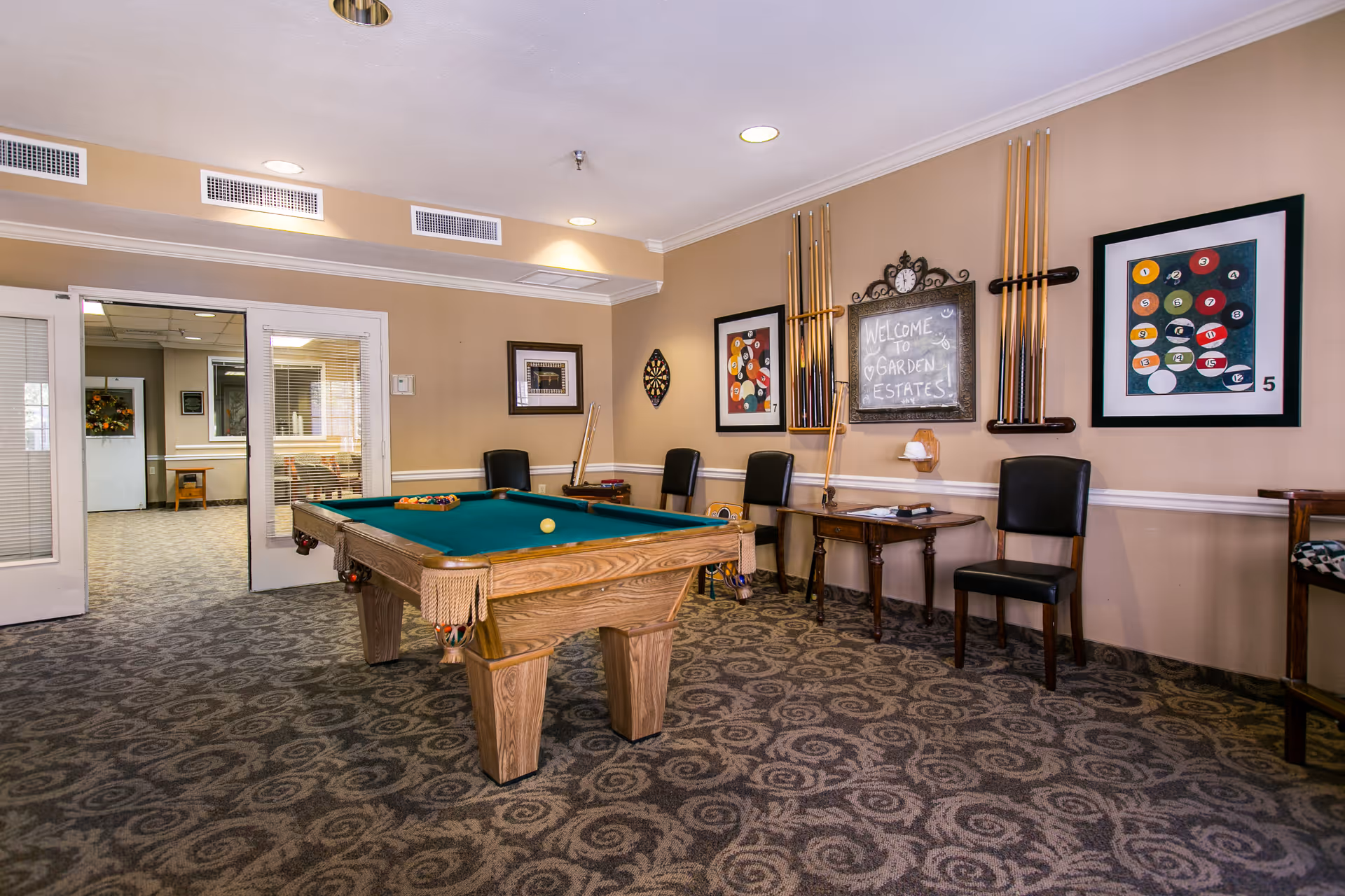 Interior view of a recreational room at Garden Estates of Corpus Christi featuring a pool table with balls arranged on it, several chairs along the wall, a dartboard, and framed pictures of pool balls. A chalkboard on the wall reads 'Welcome to Garden Estates!'. The room has patterned carpet and beige walls.