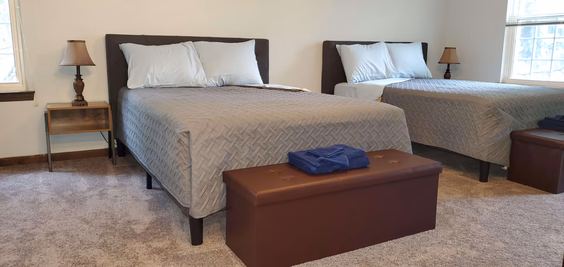 A bedroom with two beds, each with gray quilted bedspreads and white pillows. There are two brown ottomans at the foot of each bed, with folded blue towels on one ottoman. Two bedside tables with lamps are positioned next to each bed. The room has beige carpet and two windows with blinds.