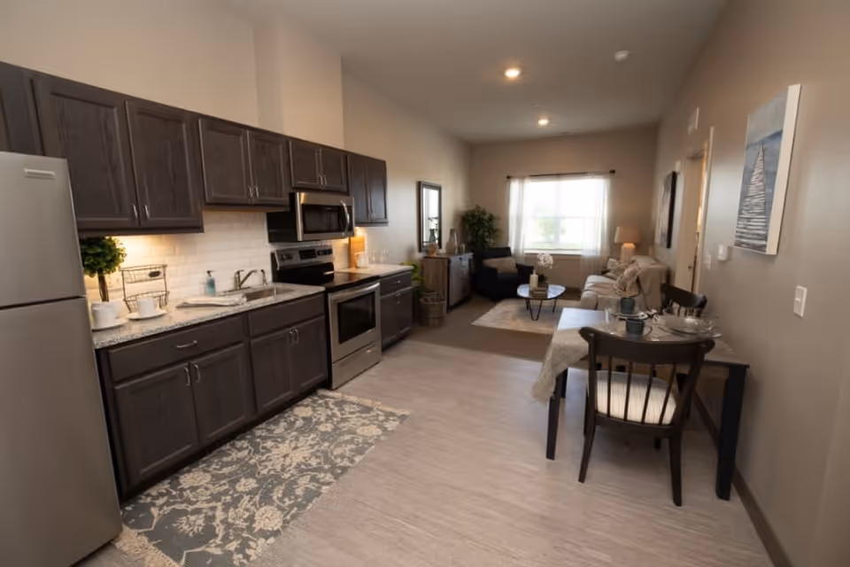 Interior view of a senior living apartment featuring a modern kitchen with dark wood cabinets, stainless steel appliances, and a patterned rug. The kitchen opens into a cozy living area with a sofa, armchair, coffee table, and a window with sheer curtains. A small dining table with two chairs is positioned along the right wall, and neutral tones decorate the space.