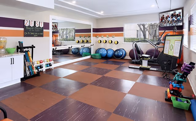 A brightly lit fitness room with checkered brown and purple flooring, exercise equipment including stability balls, dumbbells, resistance bands, and a stationary bike. There is a large mirror on one wall and a window letting in natural light. A chalkboard on the wall reads 'ZEST Fitness Challenge of the Week'.