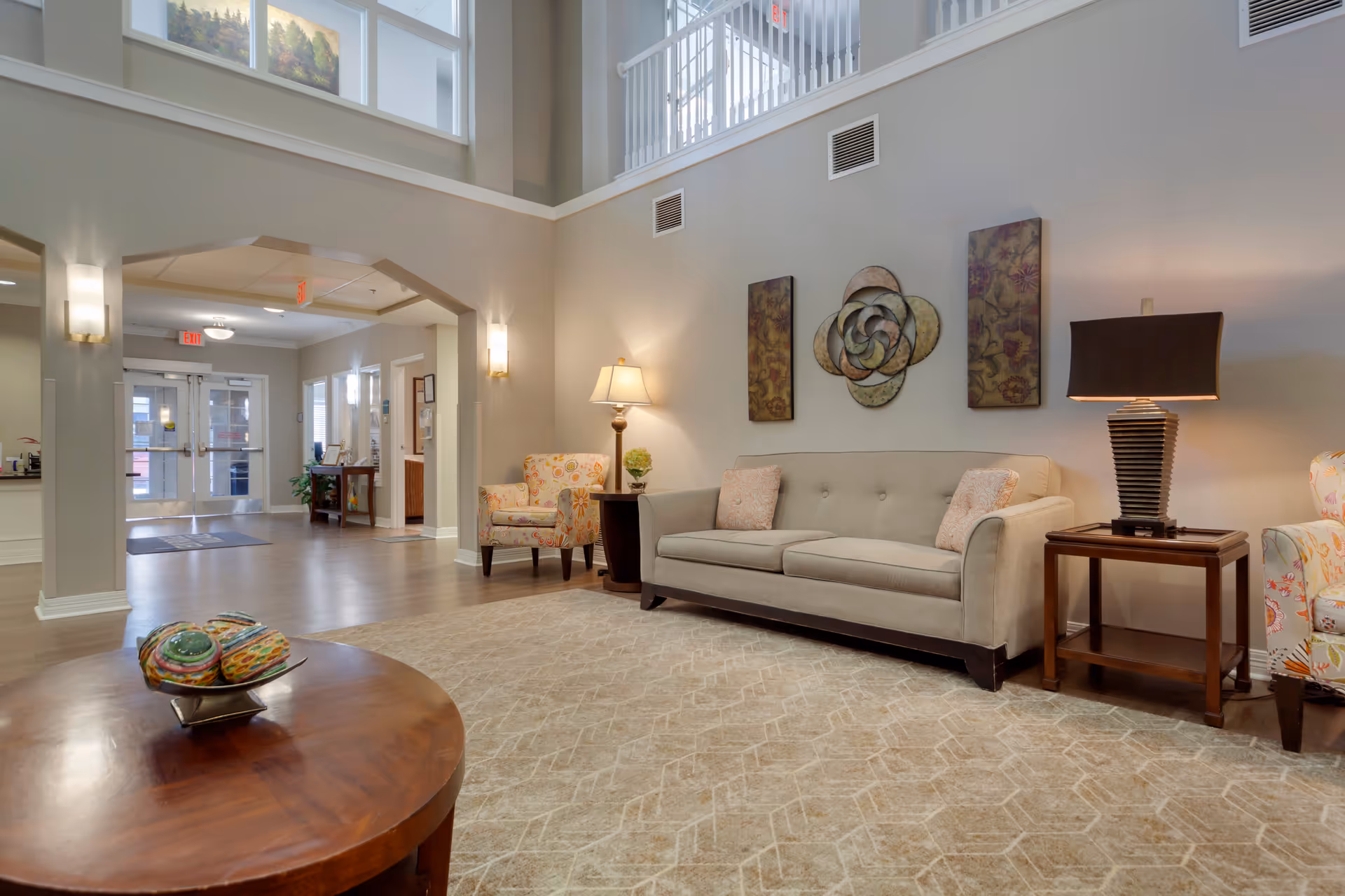 A spacious and well-lit living room area in an assisted living facility featuring a beige sofa with two patterned pillows, two floral armchairs, a wooden coffee table with decorative items, two side tables with lamps, and wall art. The room has high ceilings with windows near the top and an open archway leading to a hallway with double glass doors and exit signs.