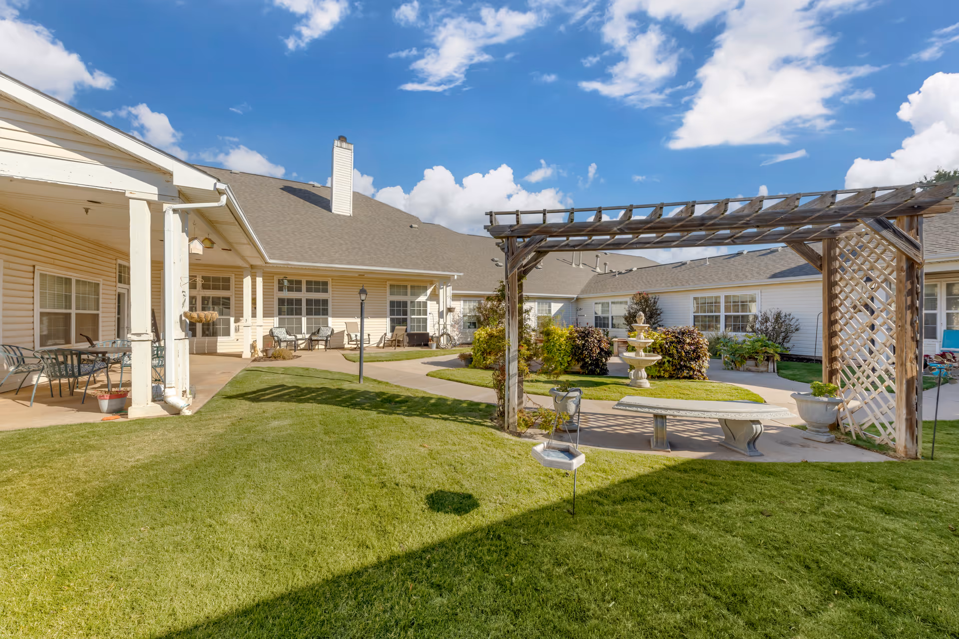 Sunlit courtyard of a senior living facility showing a wooden pergola, stone bench, fountain, patio seating and a well-kept lawn.