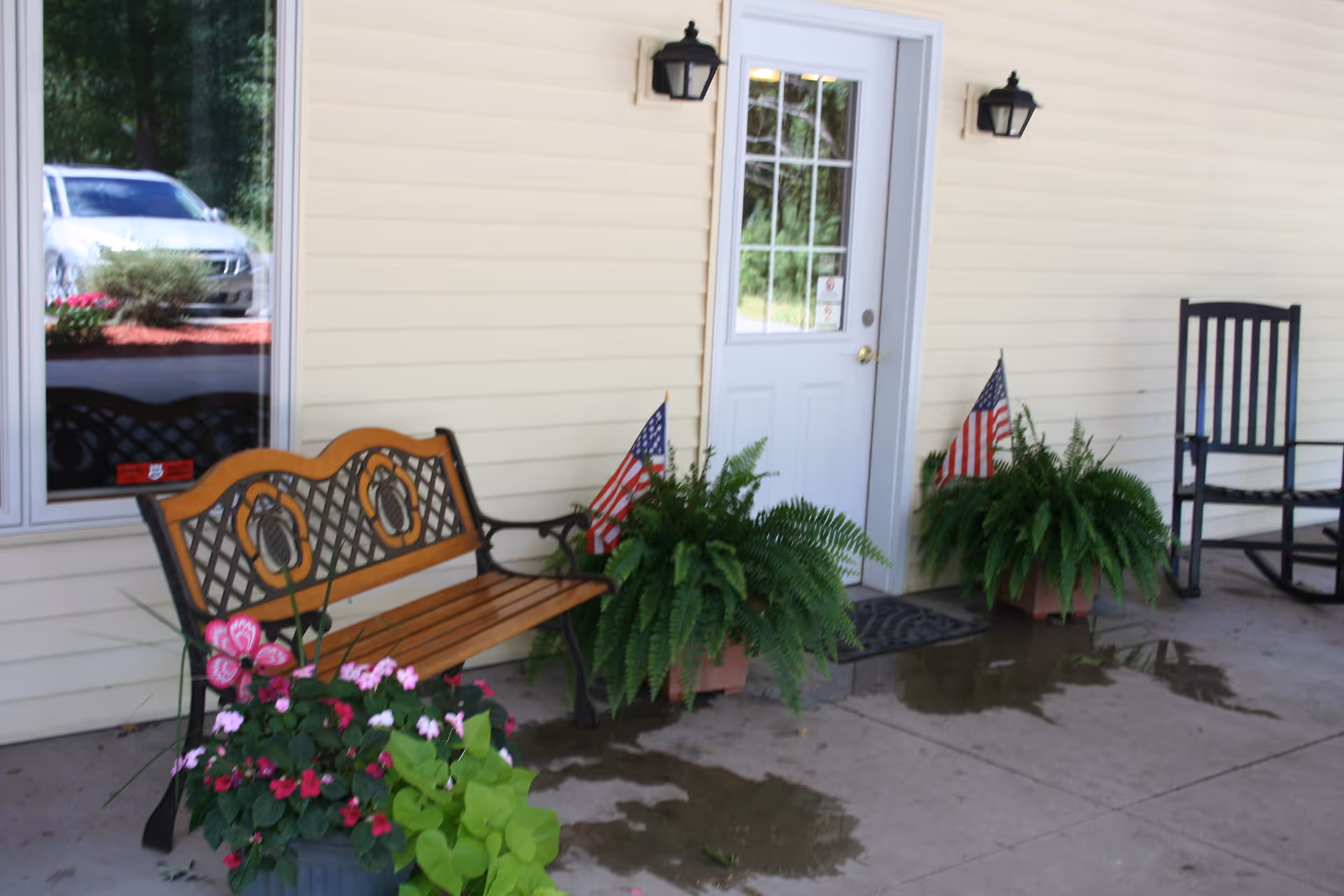 Outdoor seating area with a wooden bench, two potted ferns with small American flags, a black rocking chair, and a white door on the exterior wall of a building. There are also some pink flowers in a pot and a window reflecting a parked car.