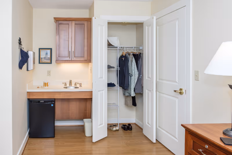 Interior view of a senior living facility room showing a small kitchenette area with a sink, a mini refrigerator, and a wooden cabinet above. Next to the kitchenette is an open closet with white double doors, containing hanging clothes and shoes on the floor. To the right, part of a wooden dresser and a lamp with a white shade are visible.