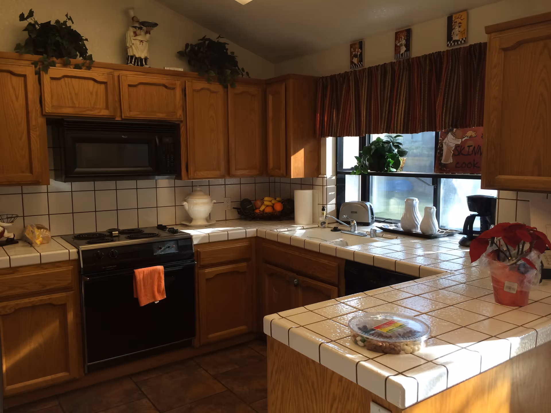 A kitchen with wooden cabinets, white tiled countertops, and a tiled backsplash. The kitchen features a black stove with an orange towel hanging on the handle, a black microwave above the stove, a double sink under a window with striped curtains, a toaster, a coffee maker, and various decorative items including plants and a poinsettia on the counter.