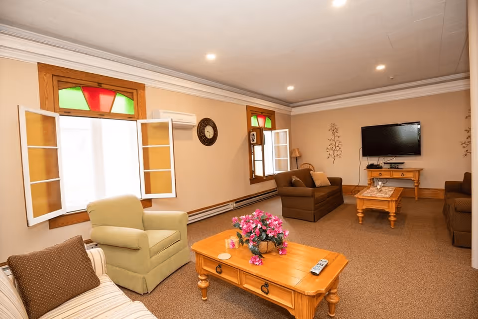 A cozy living room with beige walls and carpeted floor featuring a wooden coffee table with a pink flower arrangement, a green armchair, a striped sofa with a brown cushion, two brown sofas, a wooden side table with a lamp, a wall-mounted flat-screen TV, and two windows with stained glass accents and open white shutters.