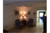 A small seating area in a hallway with two upholstered chairs and a wooden side table with a lamp. Two framed pictures hang on the wall above the chairs. The hallway leads to another room with more furniture visible in the background.