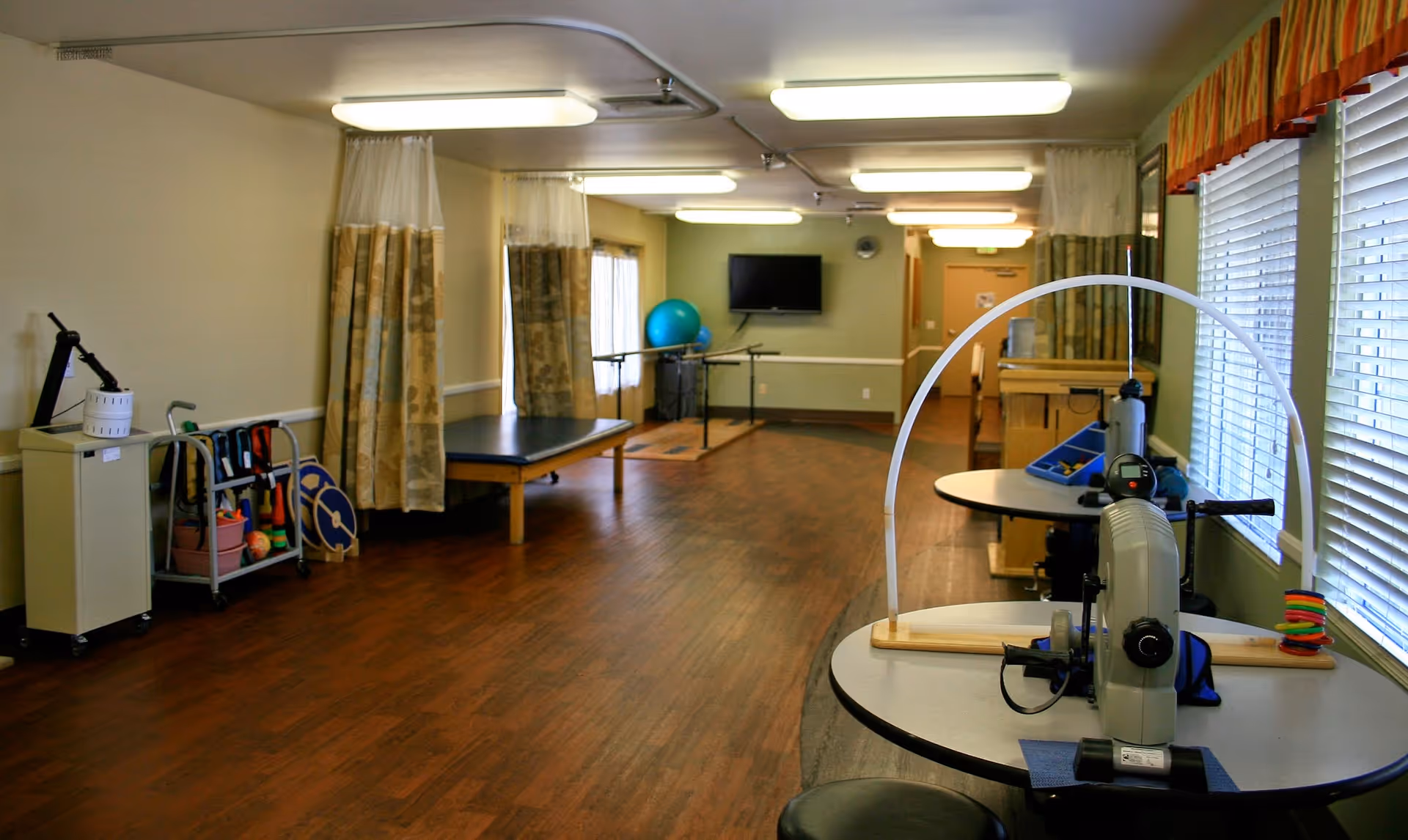 A therapy or rehabilitation room with wooden flooring, exercise and therapy equipment including a table with a hand exercise machine, a therapy bed, a large exercise ball, and various therapy tools on a cart. The room has beige walls, fluorescent ceiling lights, and windows with blinds and valances.