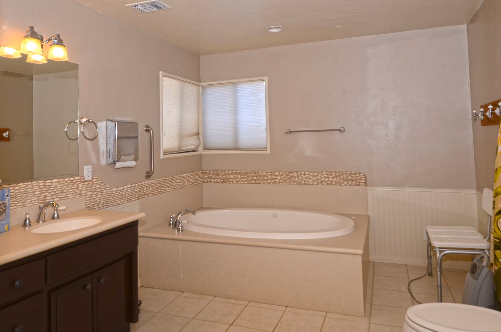 A bathroom with a large built-in bathtub, beige tiled floor, and beige walls. There is a sink with a countertop and dark brown cabinet below on the left side, a large mirror above the sink, a paper towel dispenser, and a towel ring. The bathtub has a decorative tile border around it. A window with closed blinds is above the bathtub. On the right side, there is a white shower chair, a towel rack, and a toilet partially visible.