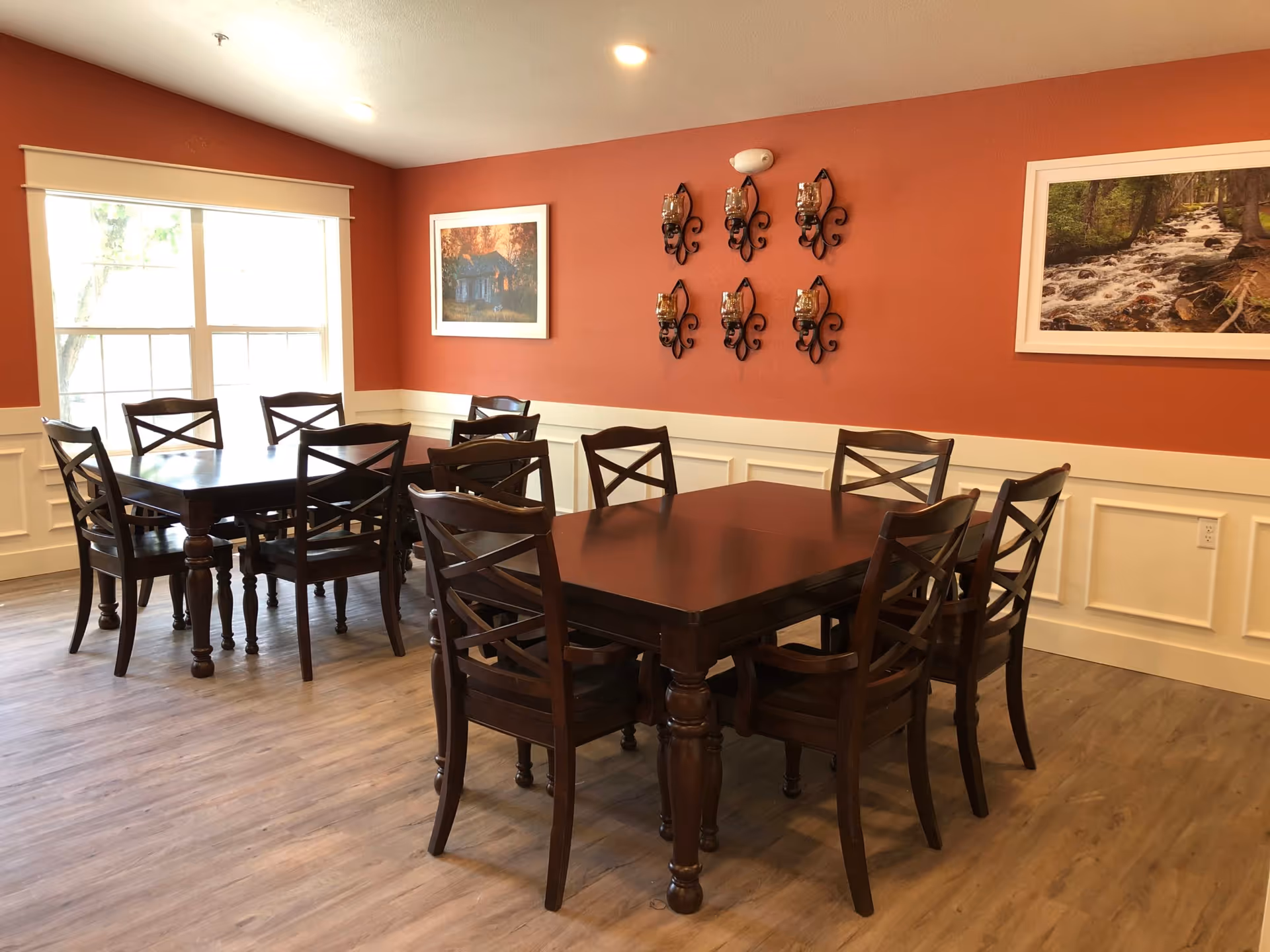 Dining area with dark wood tables and chairs, a red accent wall with artwork and decorative sconces, and a large window letting in light.