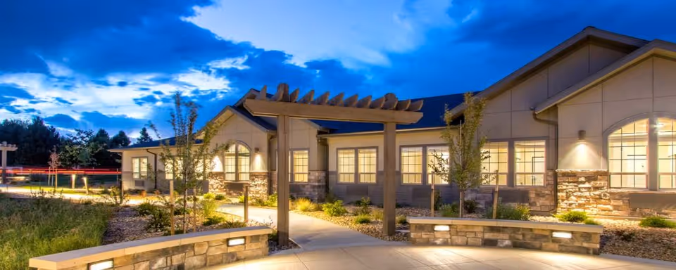 Exterior view of a single-story building at dusk with warm interior lights visible through multiple windows. The building features stone accents and a pergola over a paved walkway surrounded by landscaped greenery and small trees.