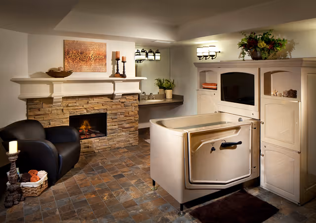 A cozy room featuring a stone fireplace with a lit fire, a black armchair, a basket with towels, a beige walk-in bathtub, a sink with a plant, and a cabinet with a TV and decorative items on top.
