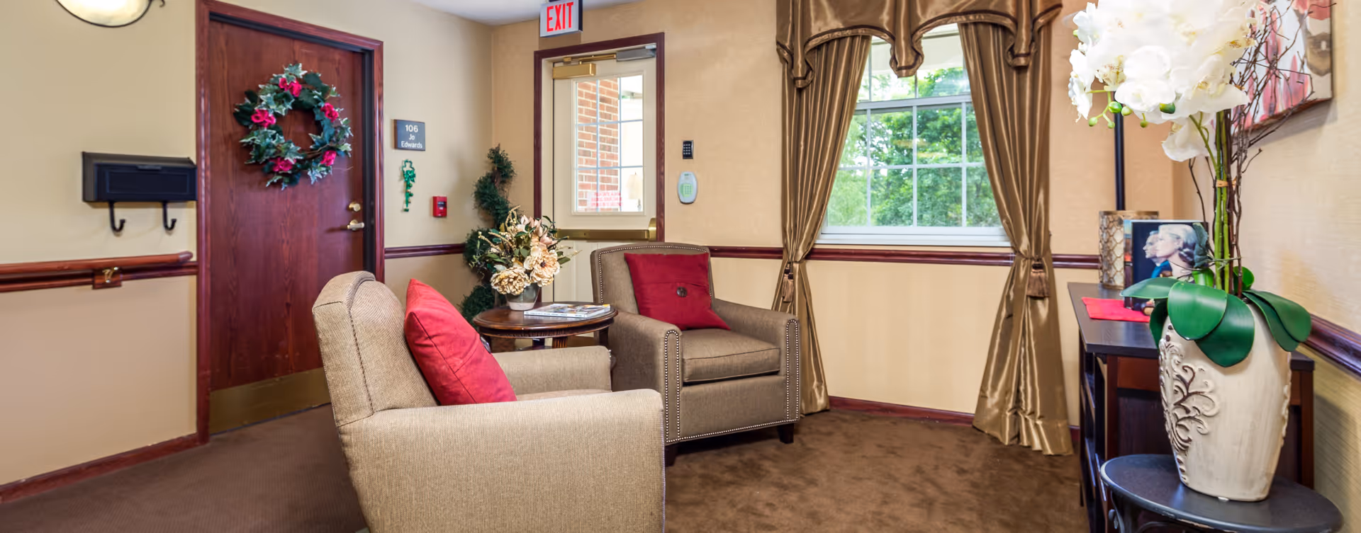 A cozy sitting area in a senior living facility with two beige armchairs featuring red cushions, a small round table with a floral arrangement, a window with gold curtains, and a door decorated with a holiday wreath. There is also a console table with a vase of white flowers and framed pictures.