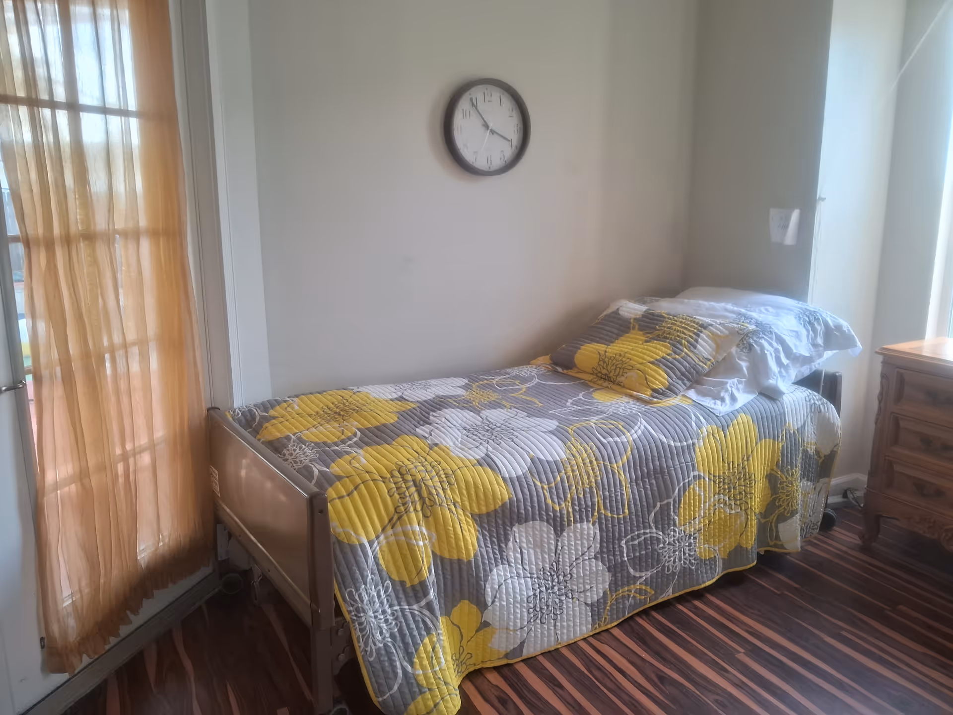 Single bed with a yellow and gray floral bedspread in a small bright bedroom with a clock on the wall.