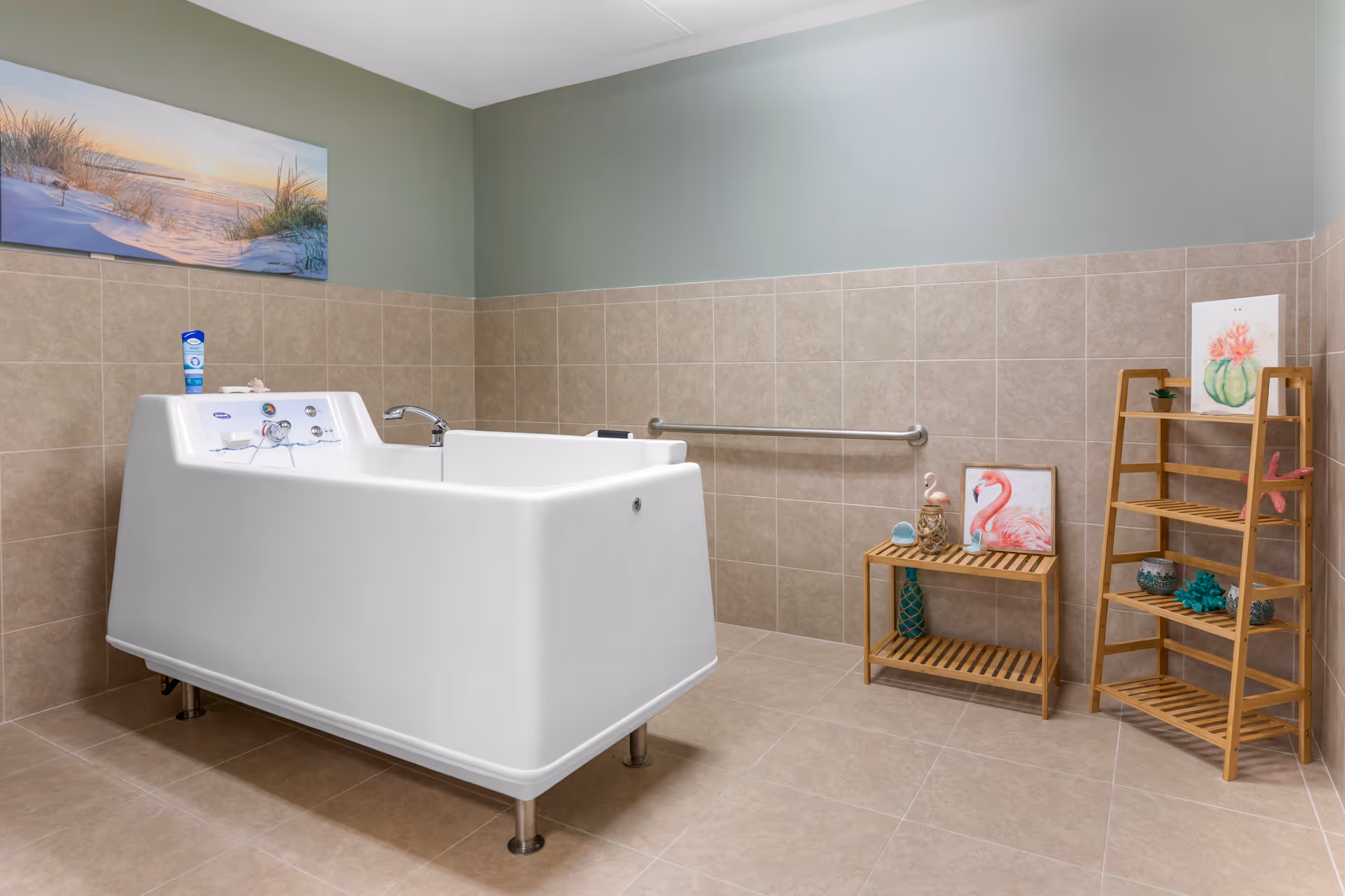 A spacious bathroom with beige tiled walls and floor, featuring a large white walk-in bathtub with controls and a faucet. On the right side, there are two wooden shelves decorated with small plants, a flamingo figurine, and framed pictures including a flamingo and a cactus. A metal grab bar is mounted on the wall behind the bathtub. Above the tiles, the wall is painted light green and a large beach-themed picture hangs on the left side.