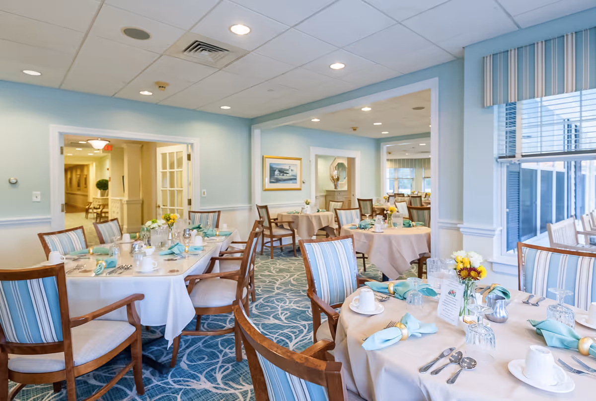 A bright and elegant dining room with multiple round and rectangular tables set with white tablecloths, turquoise napkins, silverware, glassware, and small floral centerpieces. The room features light blue walls, large windows with striped valances, and wooden chairs with striped cushions. The space is well-lit with recessed ceiling lights and has a patterned blue carpet.