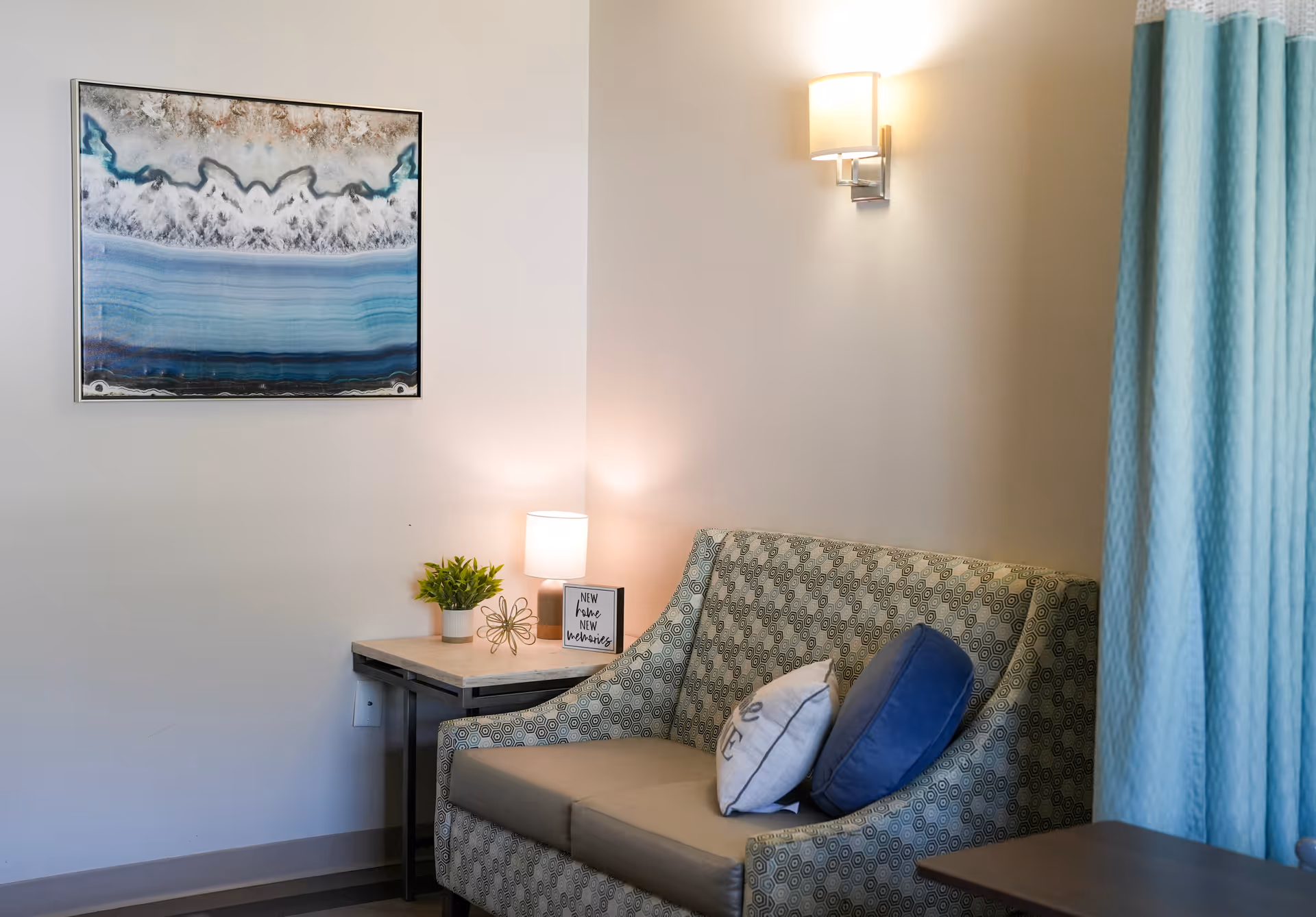 Small sitting area with a patterned loveseat, side table holding a lamp and plant, abstract wall art, and a blue curtain.