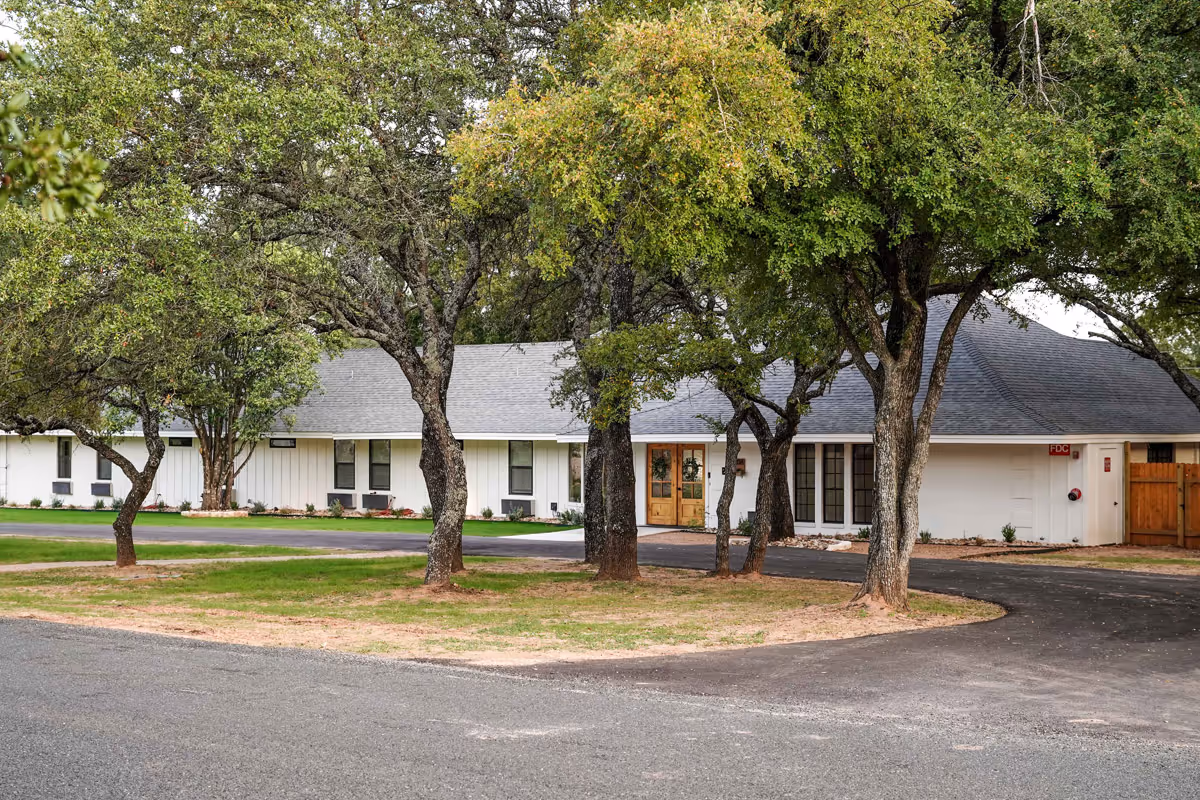 Exterior view of a single-story assisted living facility building with white walls and a gray roof, partially obscured by several large trees in the foreground. The building has multiple windows and a wooden double-door entrance. The surrounding area includes a paved driveway and patches of grass.