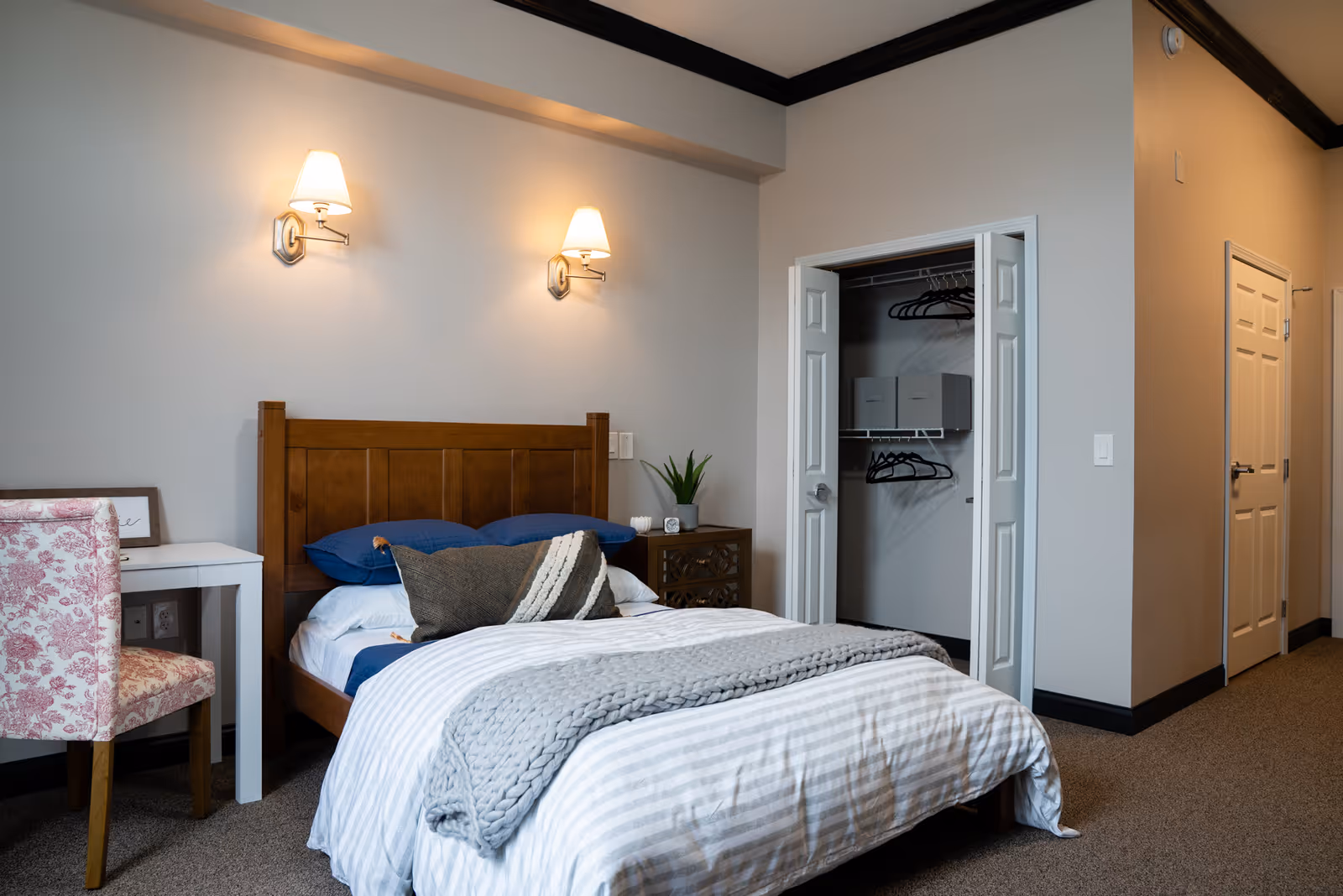A neatly made bed with striped bedding and a knitted throw blanket in a bedroom. The bed has a wooden headboard and is flanked by a nightstand with a small plant and decorative items. There are two wall-mounted lamps above the bed. To the left, there is a white desk with a pink patterned chair. An open closet with hangers and storage boxes is visible on the right side of the room.