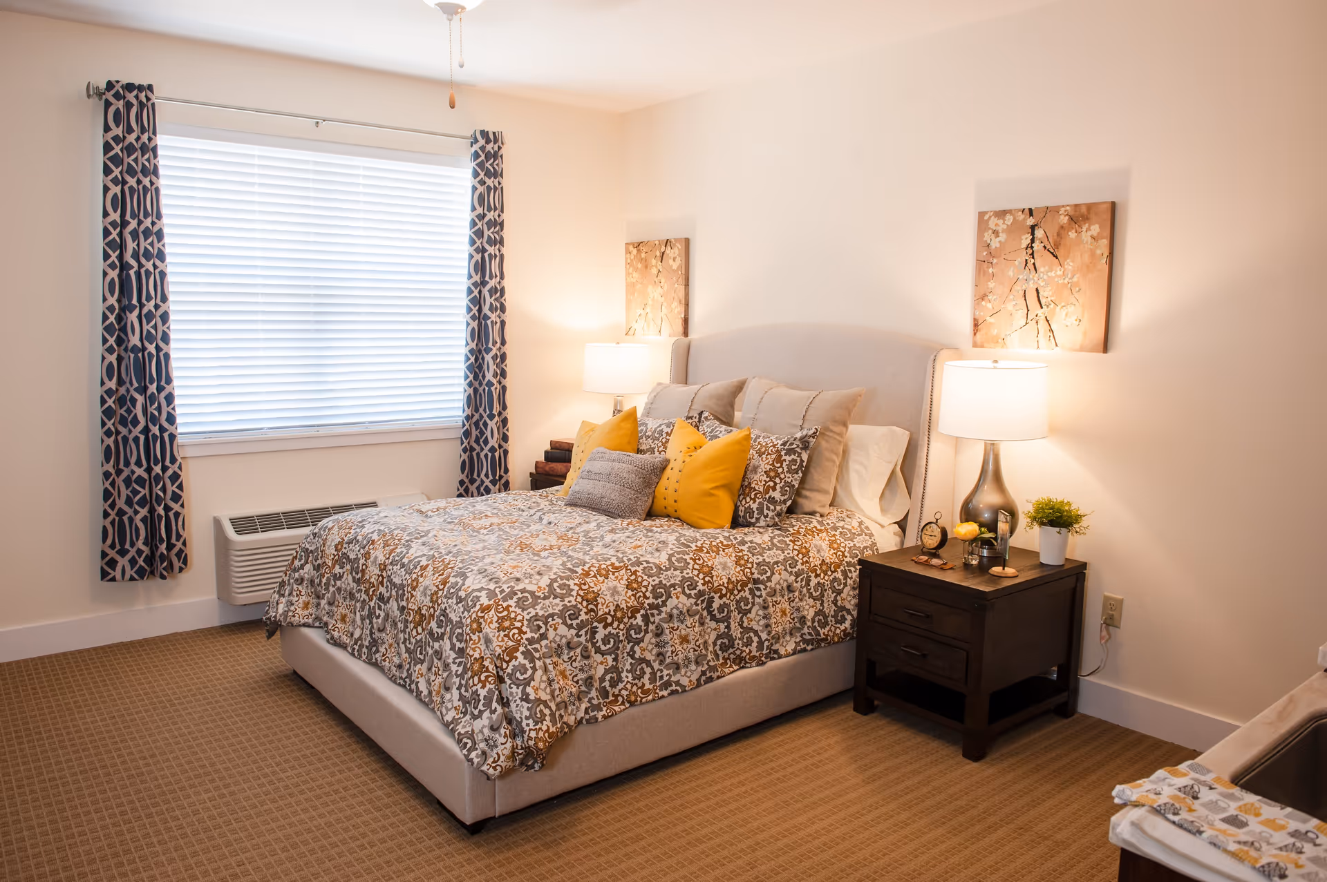 A cozy bedroom with a large bed featuring a patterned bedspread and multiple pillows in shades of beige, yellow, and gray. There are two bedside tables with lamps, decorative items, and a small plant. A window with patterned curtains and blinds is on the left wall, and two floral paintings hang above the bed.