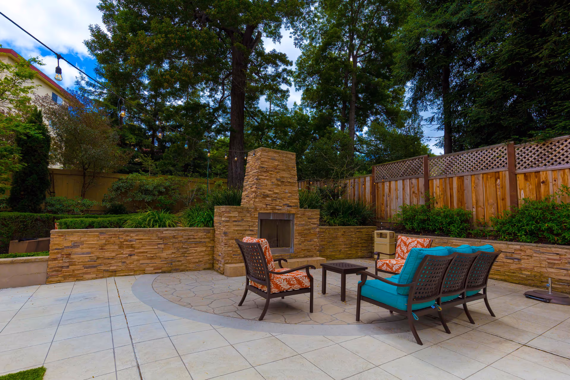 Outdoor patio with a stone fireplace, cushioned chairs and a small table, string lights, and trees with a wooden fence in the background.