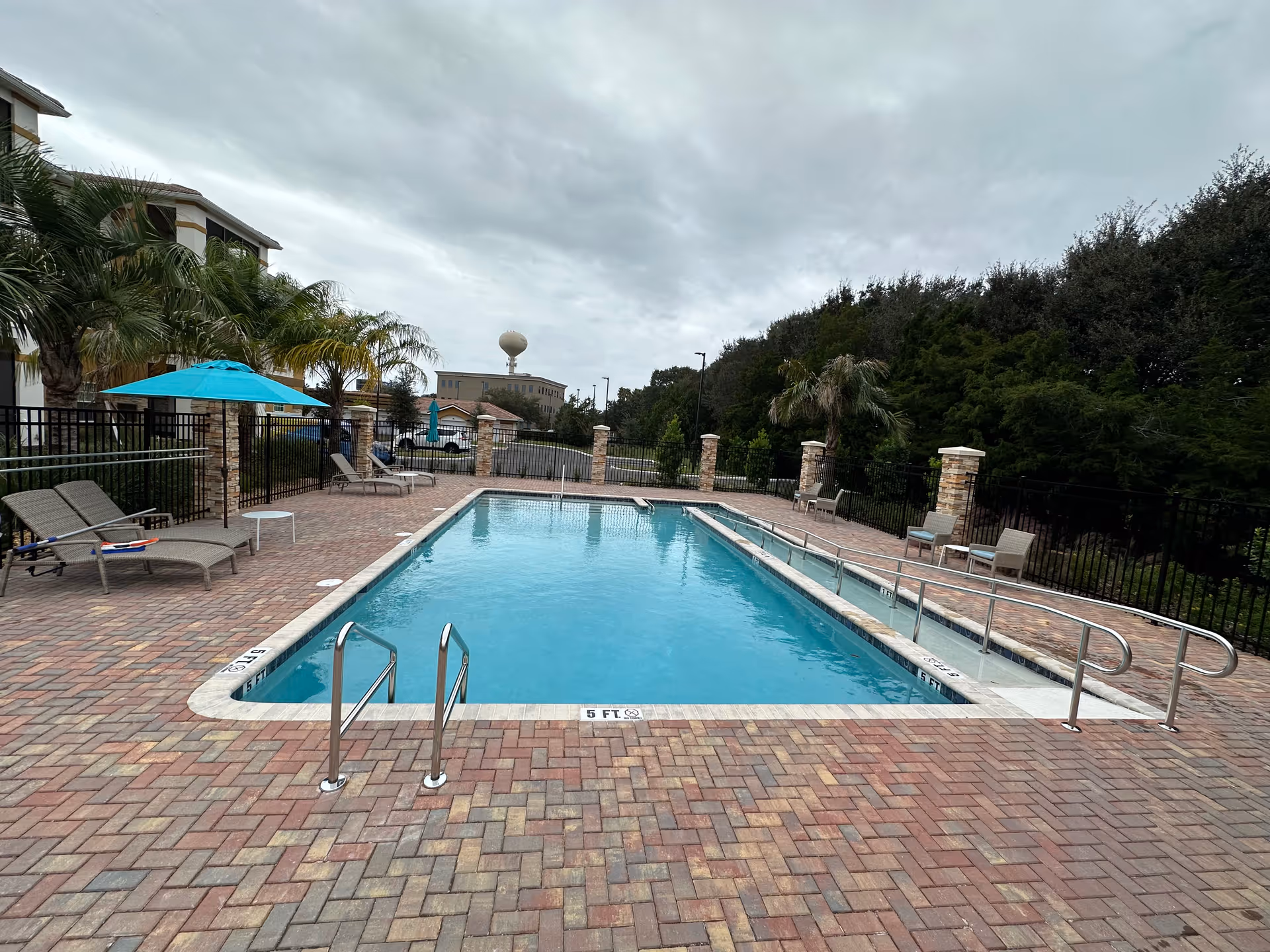 Outdoor swimming pool with clear blue water surrounded by a brick patio. There are lounge chairs and umbrellas on the left side, and a ramp with handrails on the right side of the pool. Palm trees and other greenery are visible around the pool area, with a cloudy sky overhead.
